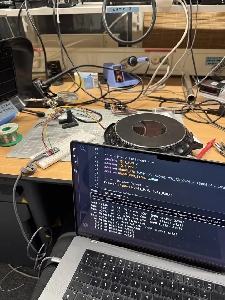 A laptop shows Arduino code defining the jog's pins and a serial monitor full of position readings; behind it the jog wheel sits next to a breadboard with a small microcontroller wired to it.