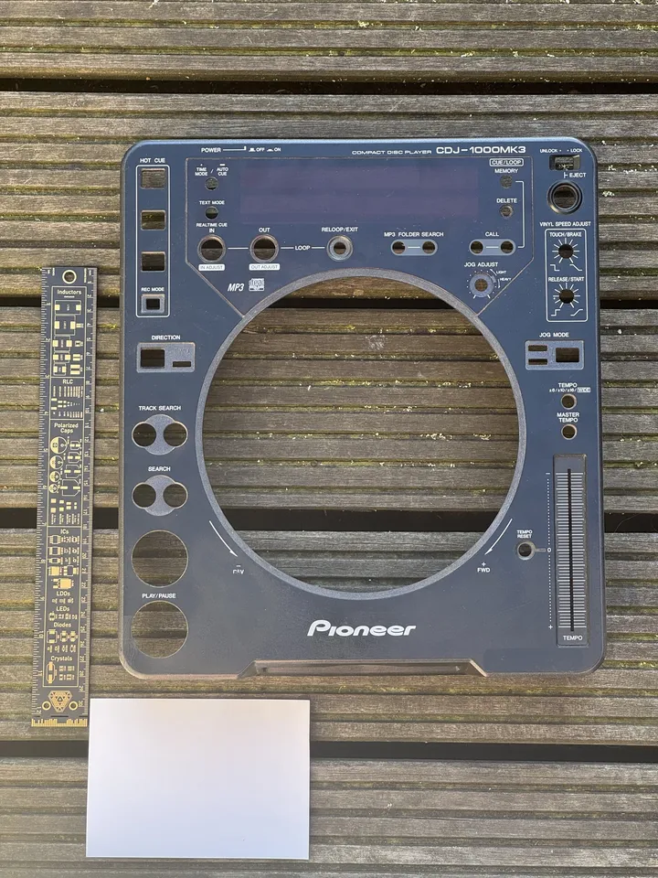 The faceplate of a Pioneer CDJ-1000MK3 on wooden decking, next to a circuit-board ruler and a white card.
