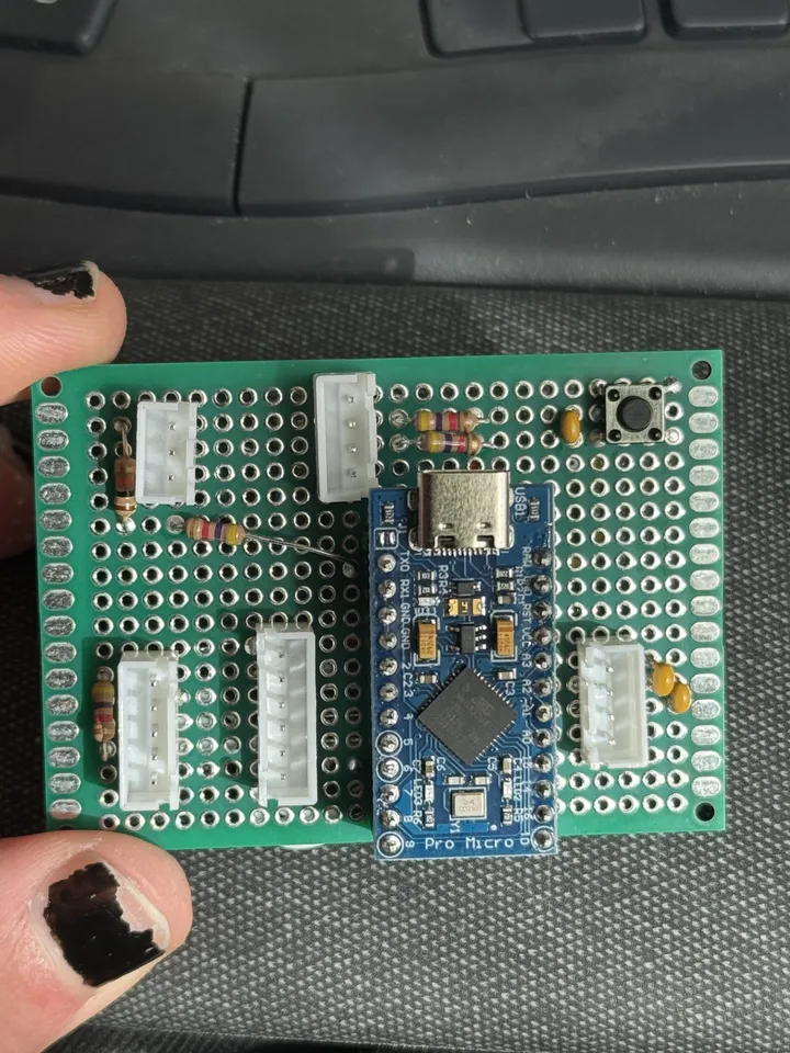 A green perfboard held in fingers, with a blue Arduino Pro Micro in the middle, white connectors around it, a few resistors and capacitors, and a small push button in a corner.