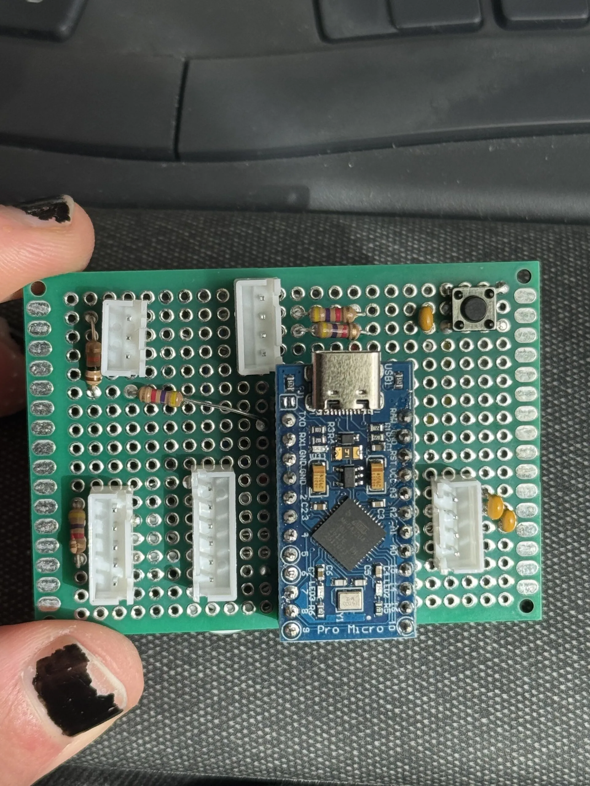 A green perfboard held in fingers, with a blue Arduino Pro Micro in the middle, white connectors around it, a few resistors and capacitors, and a small push button in a corner.