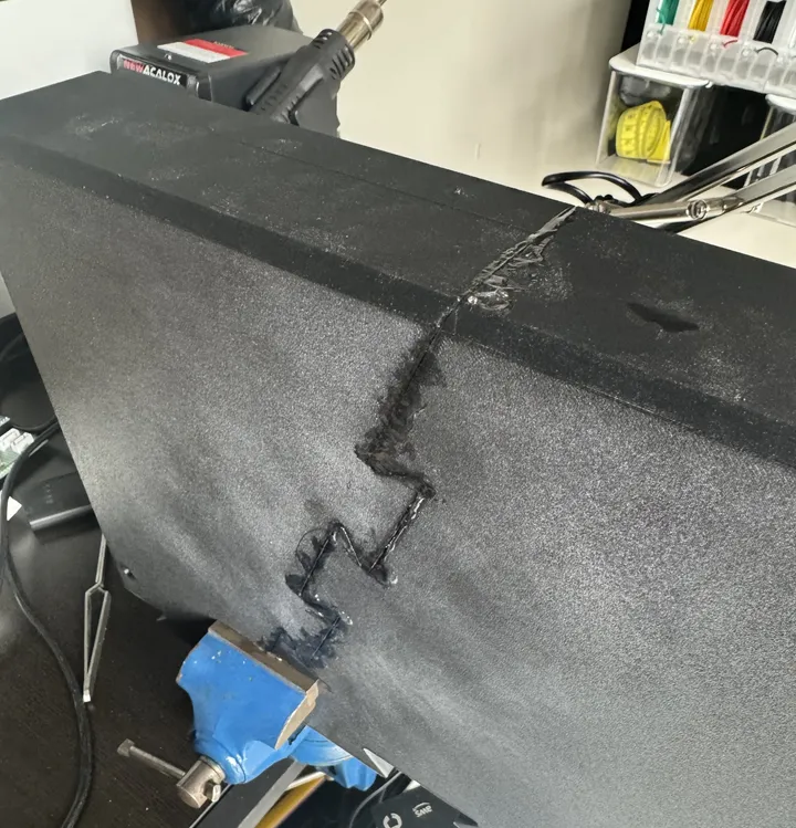 A large black textured printed panel clamped in a blue vise; a jagged, puzzle-shaped seam runs across it, filled with melted black plastic.