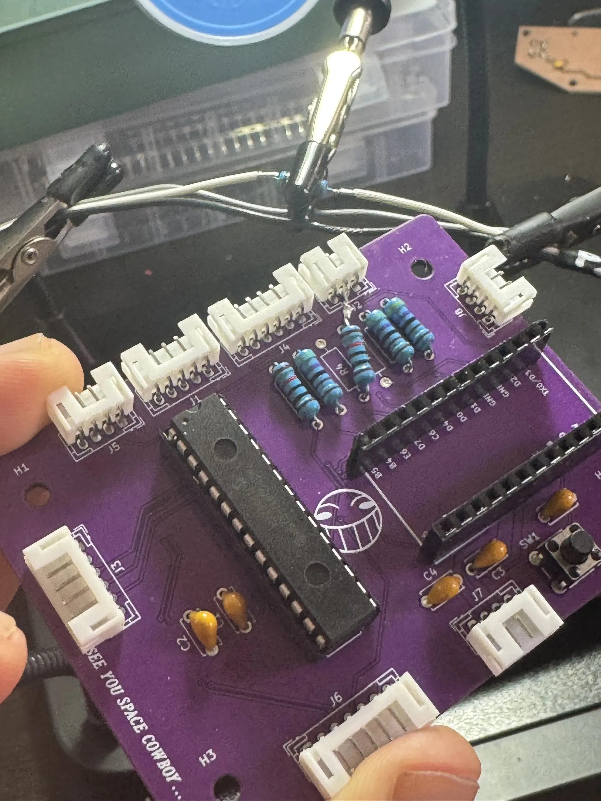 A purple board held between fingers: a long 28-pin chip, two rows of header sockets for a microcontroller, blue resistors, white connectors along its edges and a laughing-face logo.