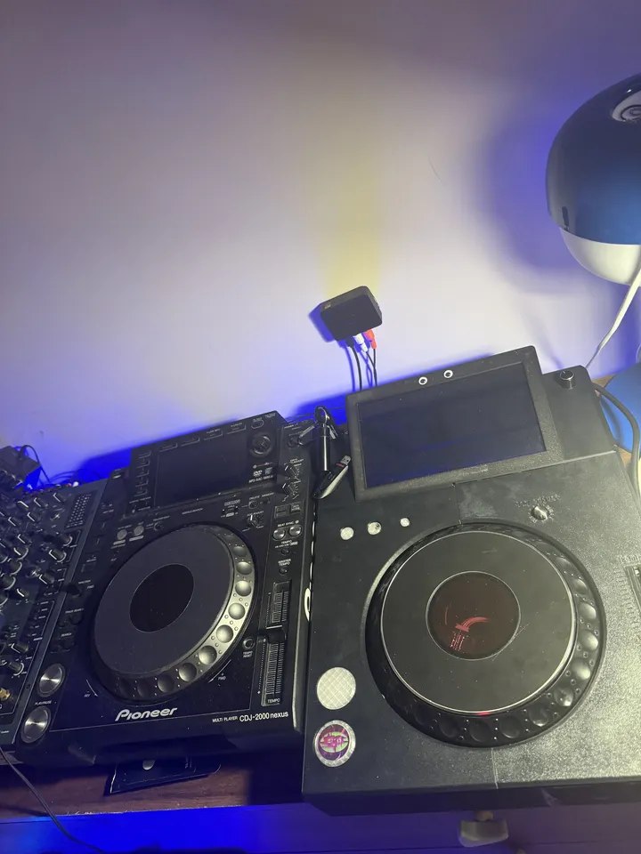 Two decks side by side under blue light: a Pioneer CDJ-2000NXS on the left, and on the right the black 3D-printed TriMixxx with the same kind of jog wheel, its screen off and two googly eyes on the screen frame.