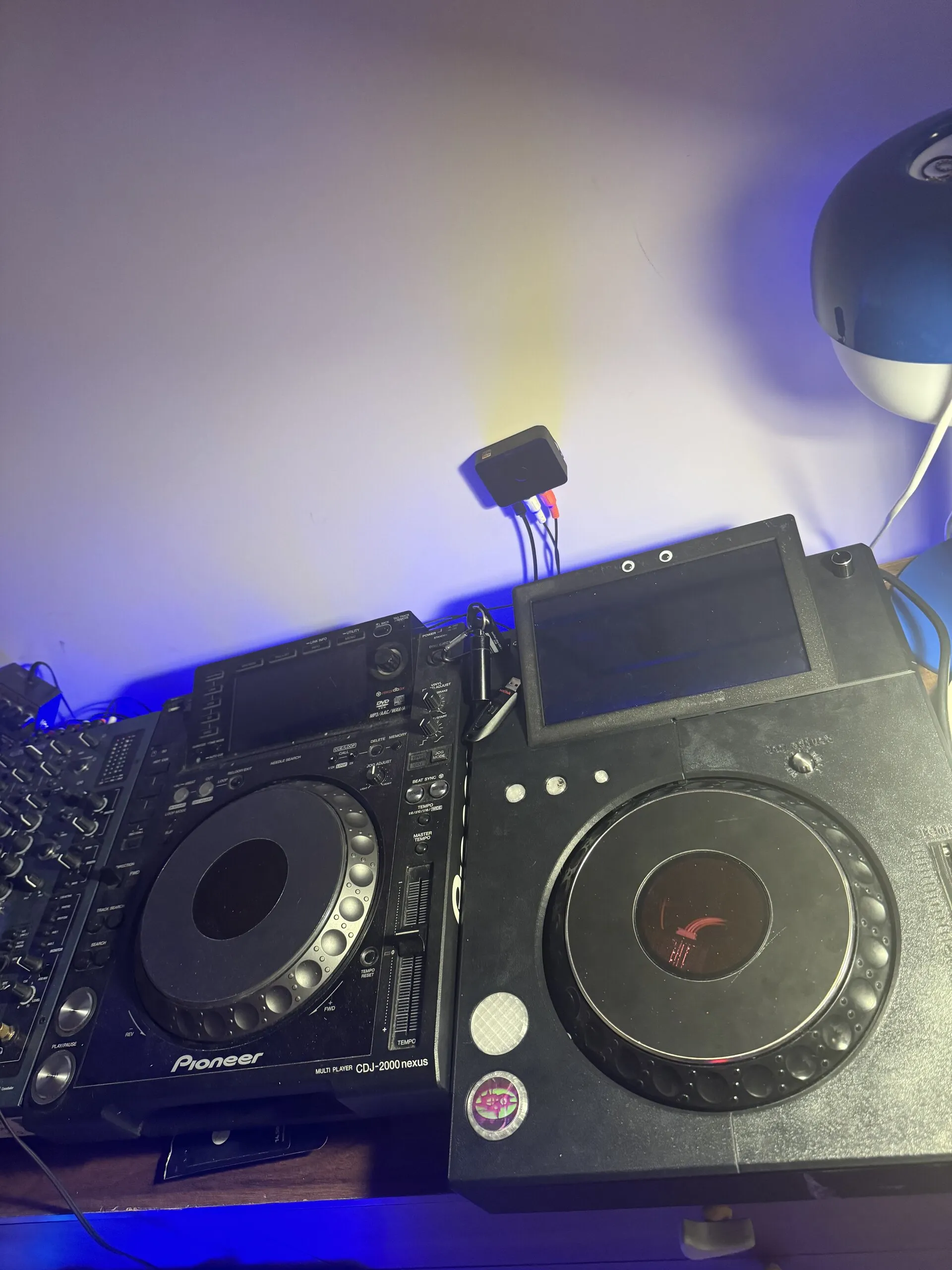 Two decks side by side under blue light: a Pioneer CDJ-2000NXS on the left, and on the right the black 3D-printed TriMixxx with the same kind of jog wheel, its screen off and two googly eyes on the screen frame.