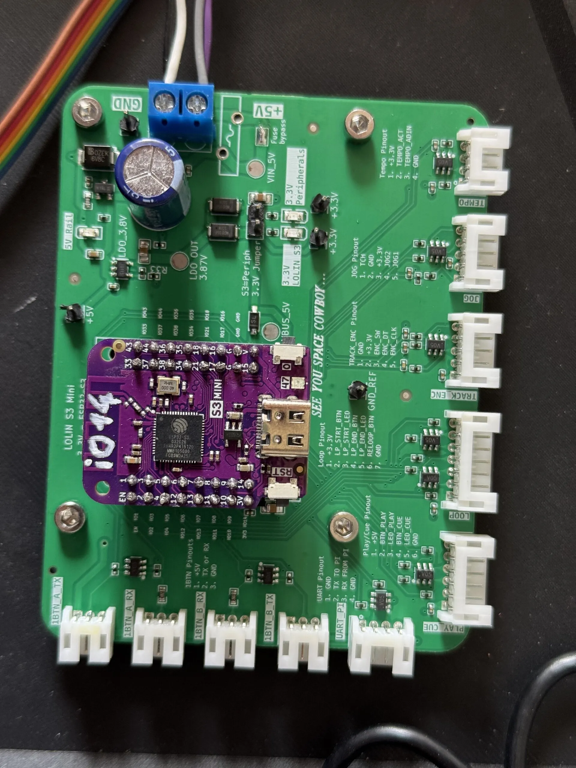 A green board with a purple ESP32-S3 module in the centre, a blue screw terminal for 5 V, and white connectors along two edges labelled for the two rings, the Pi's UART, the tempo fader, the jog, the encoder, and the loop and play/cue boards.