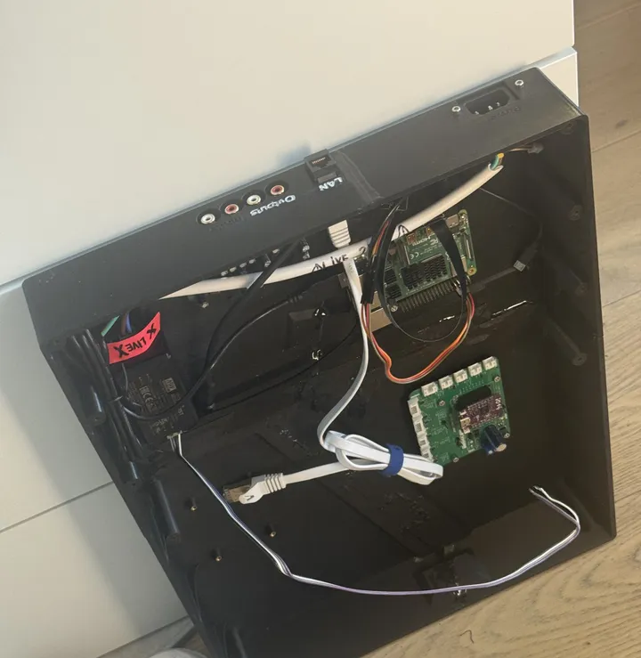 Looking into an open black printed tray: a Raspberry Pi 4, the green controller board with its purple module, a power supply, and cables to a rear panel with RCA outputs, a network port and a mains inlet.