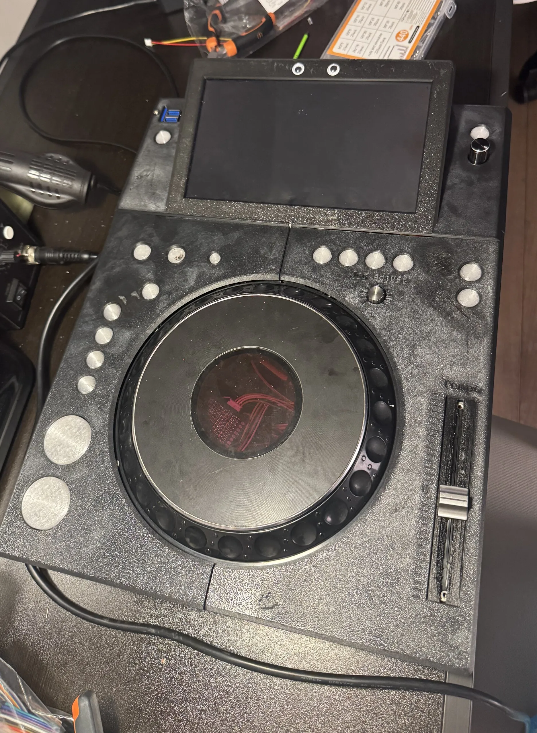 An all-black 3D-printed deck on a workbench: a screen in a raised frame with two googly eyes stuck on top, round silver buttons around a CDJ jog wheel whose centre shows red, a tempo fader, and large round CUE and PLAY buttons.