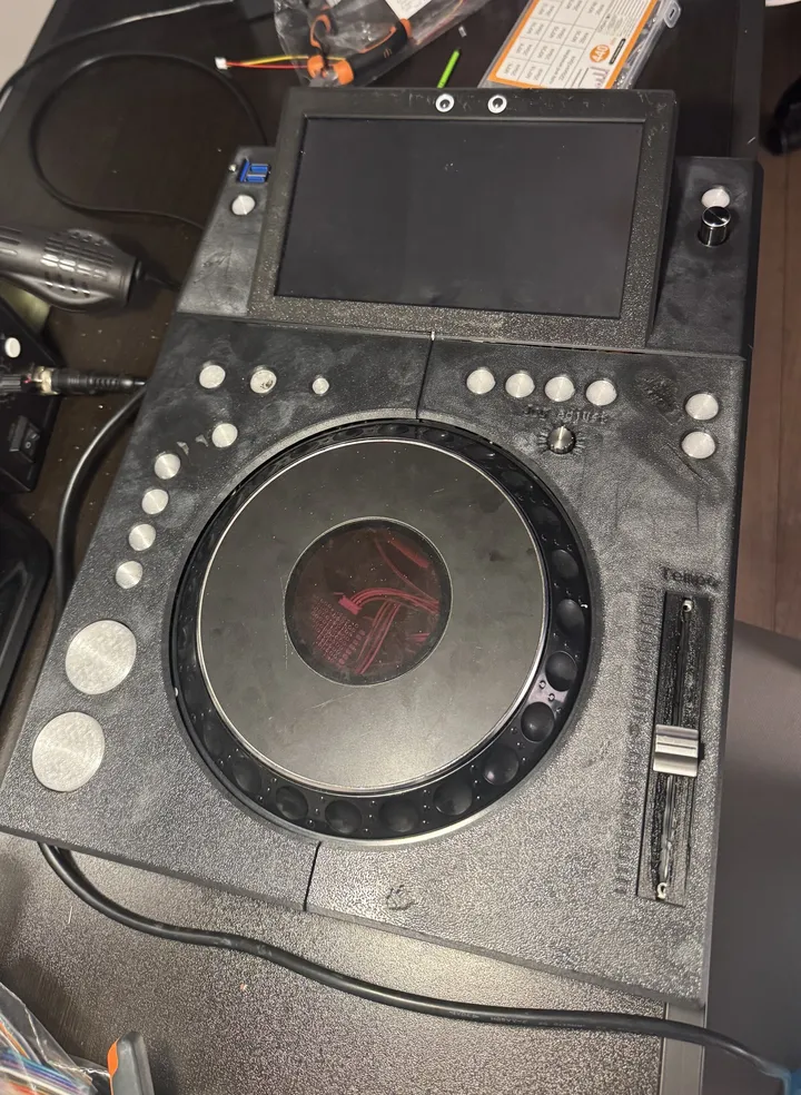 An all-black 3D-printed deck on a workbench: a screen in a raised frame with two googly eyes stuck on top, round silver buttons around a CDJ jog wheel whose centre shows red, a tempo fader, and large round CUE and PLAY buttons.