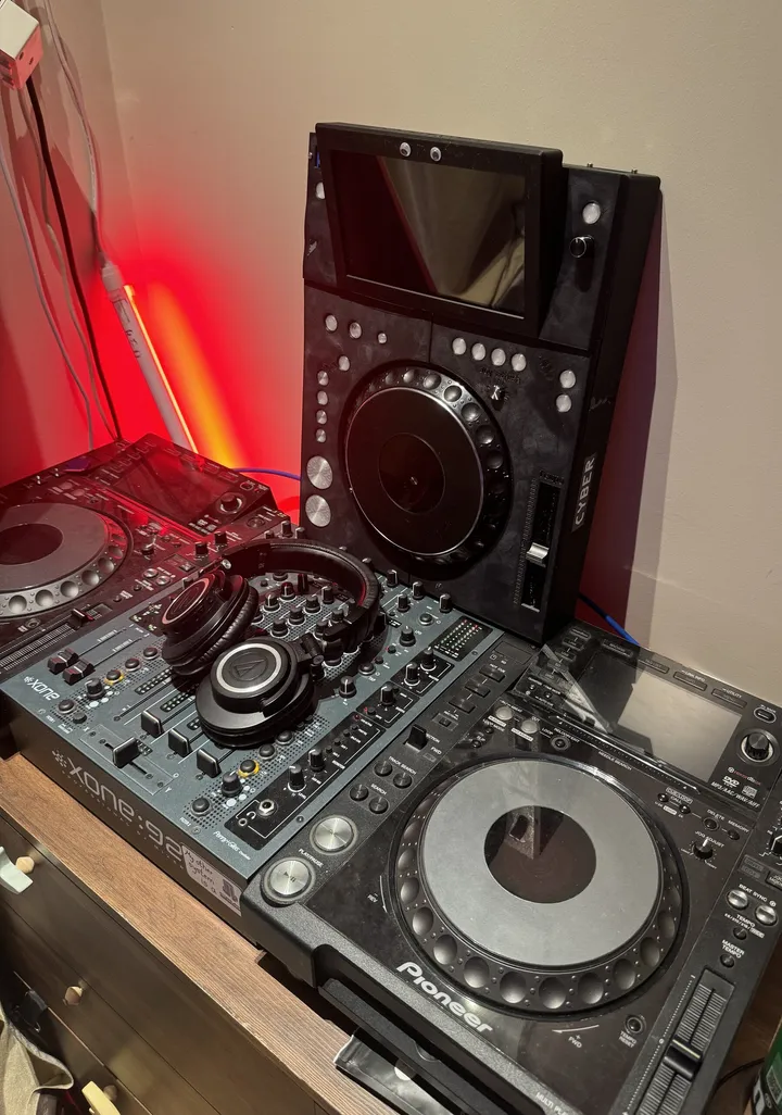 Two Pioneer CDJ-2000NXS players either side of an Allen & Heath Xone:92 mixer with headphones on it; the black TriMixxx deck stands upright against the wall behind them under a red LED strip.