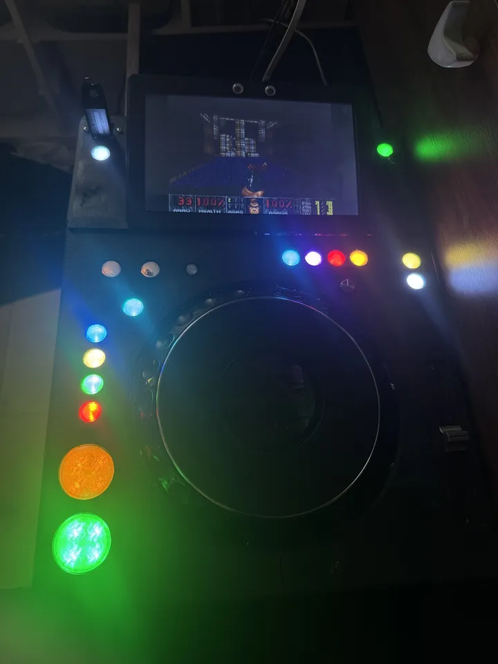 The deck in a dark room, its screen showing DOOM's first level and status bar; the buttons around the jog glow blue, purple, red, yellow and white, the big CUE button orange and PLAY green.