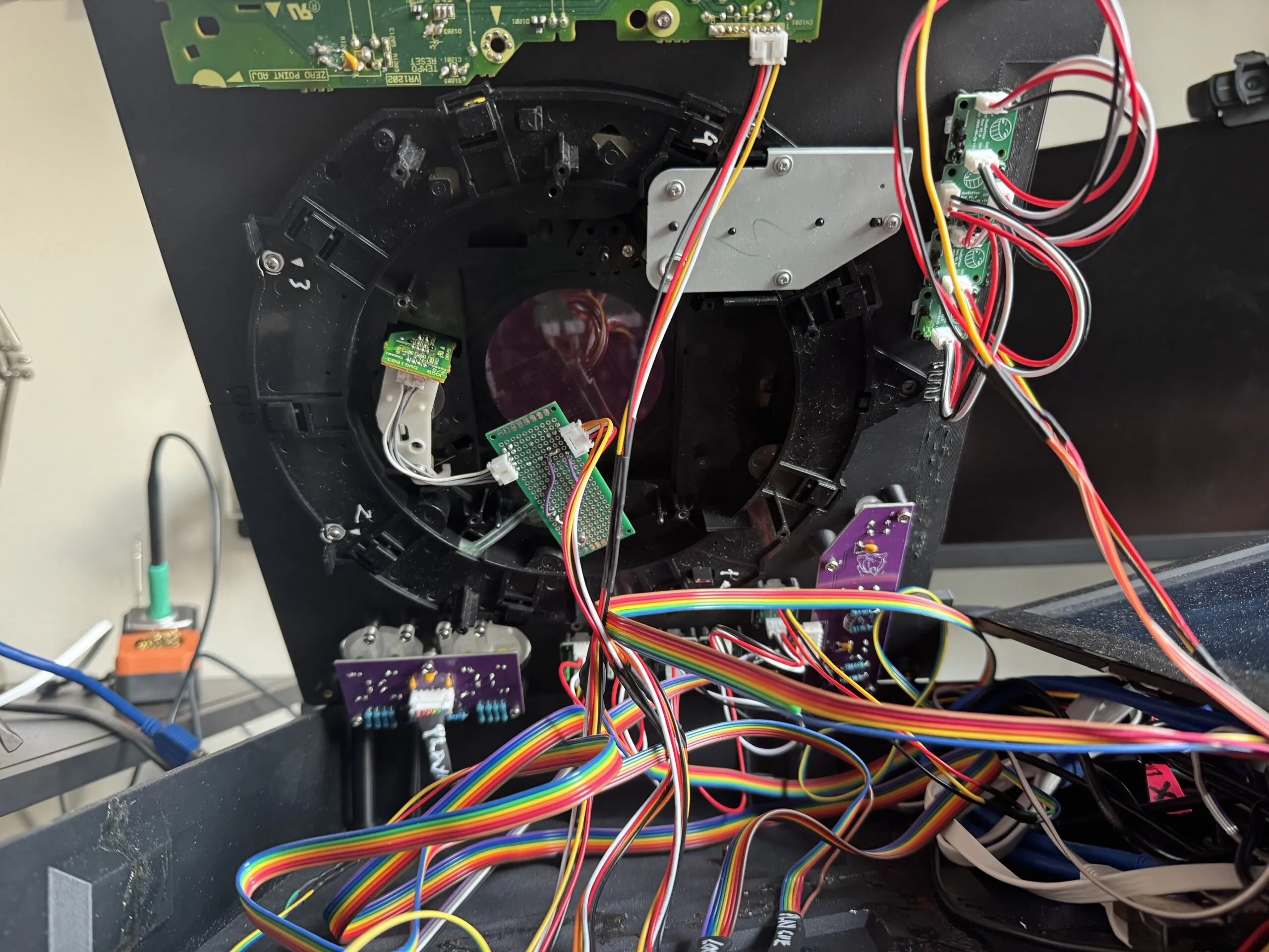 The underside of the top plate, opened like a lid: the black frame and metal bracket of a CDJ jog wheel with a small sensor board, a column of small green button boards linked by red and white wires, two purple boards, and bundles of rainbow ribbon cable.