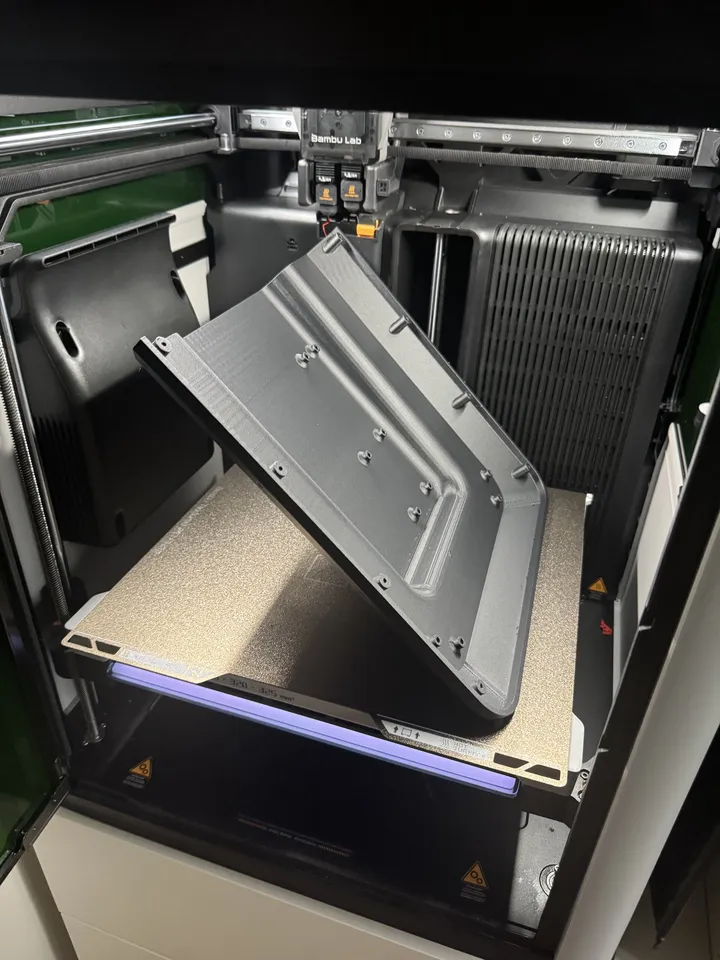 Inside a 3D printer, a large black ribbed panel with screw bosses stands at an angle on the textured build plate, the toolhead above it.
