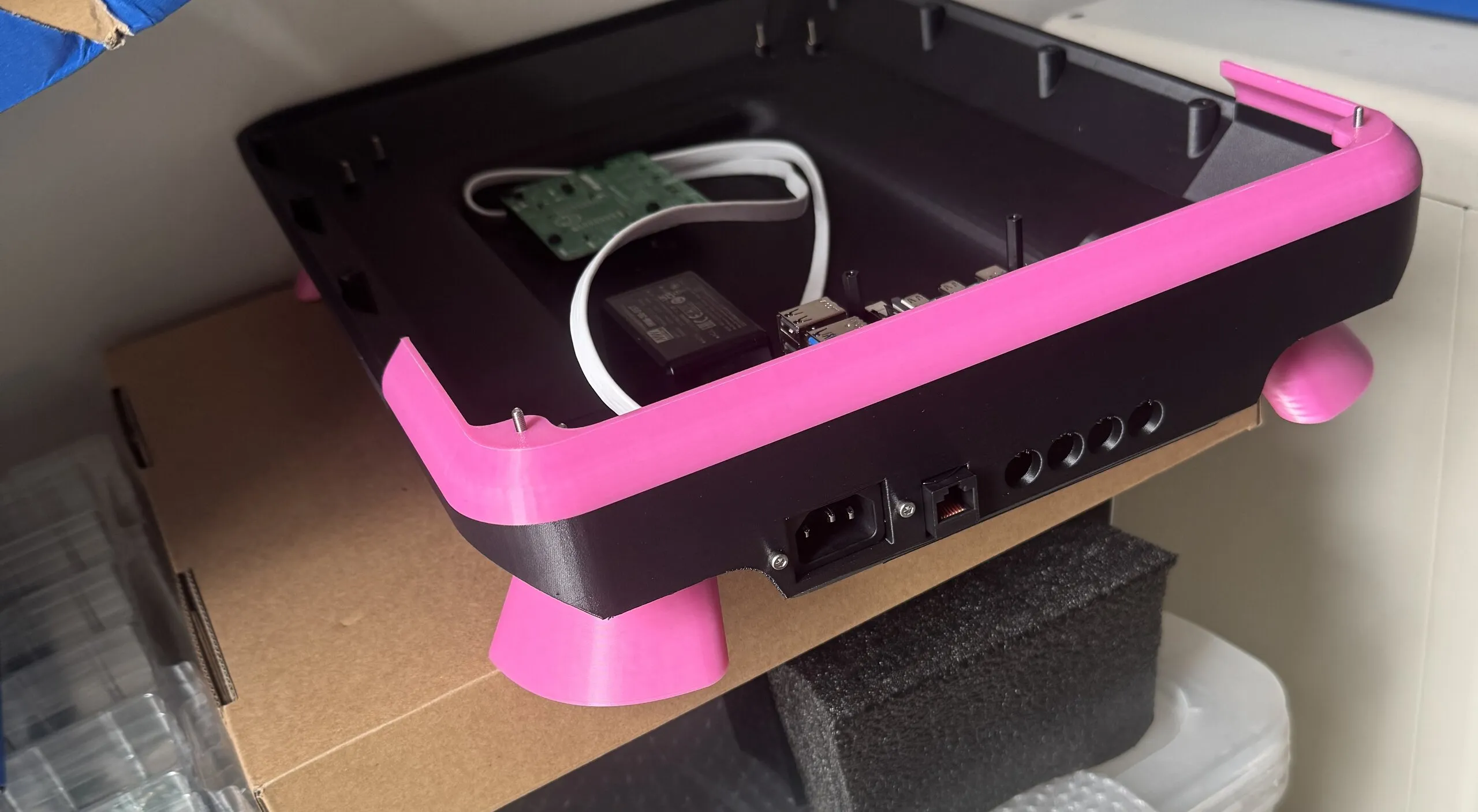 A black printed tray with pink corner bumpers and pink cone-shaped legs on a shelf; its rear panel has a mains inlet, an Ethernet socket and a row of round jack holes, and a Raspberry Pi and a power supply sit inside.
