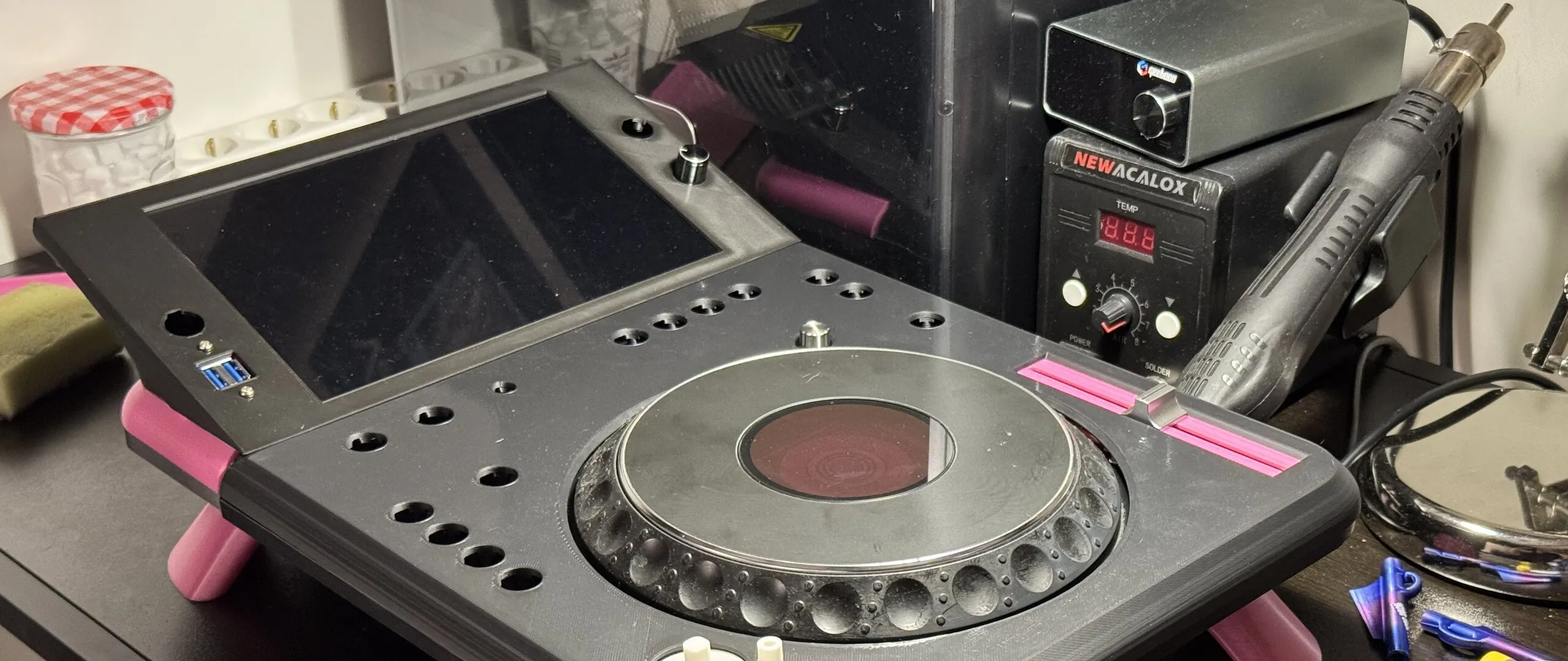 A black 3D-printed deck at an angle on a desk: a large tilted screen, empty round holes where buttons will go, a CDJ jog wheel, a pink inlay around the tempo fader slot and pink cone-shaped legs; a soldering station and a hot-air gun behind.