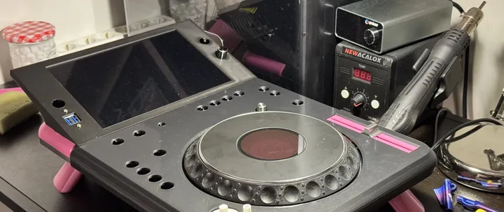 A black 3D-printed deck at an angle on a desk: a large tilted screen, empty round holes where buttons will go, a CDJ jog wheel, a pink inlay around the tempo fader slot and pink cone-shaped legs; a soldering station and a hot-air gun behind.