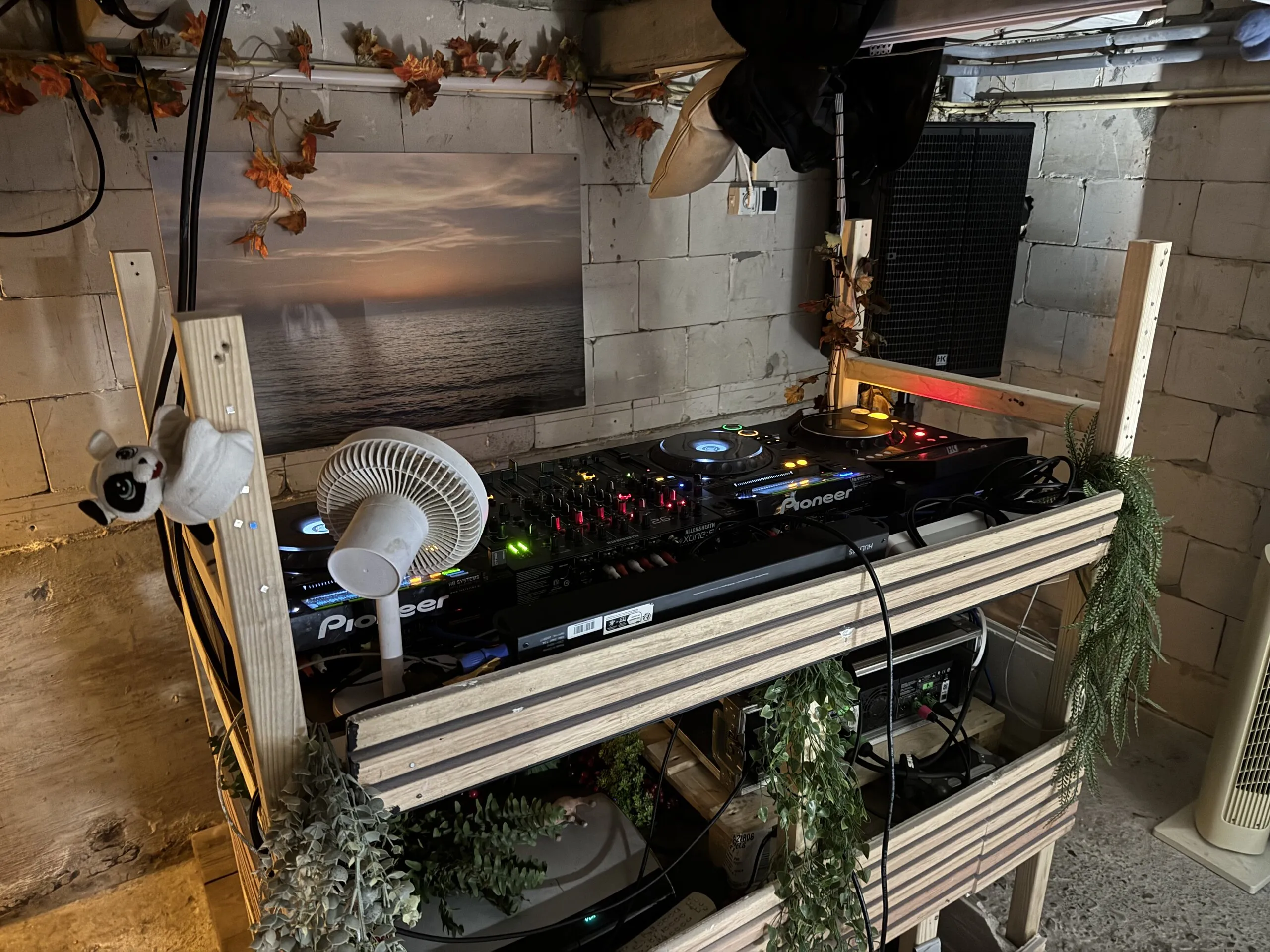 A booth of pale wooden slats with trailing plants, holding two players and a mixer, a desk fan and a small plush toy; behind it, a large photo of a sea at sunset on a concrete-block wall beside a loudspeaker.