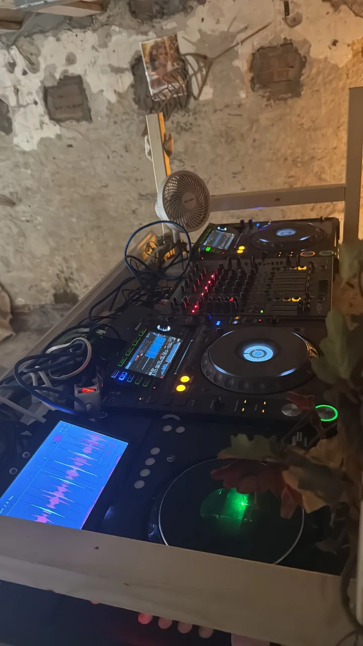A dim DJ booth against a crumbling plaster wall: the TriMixxx deck in the foreground with a pink and blue waveform on its screen and a green glow in its jog, next to another player and a mixer; a desk fan and a garland of autumn leaves above.