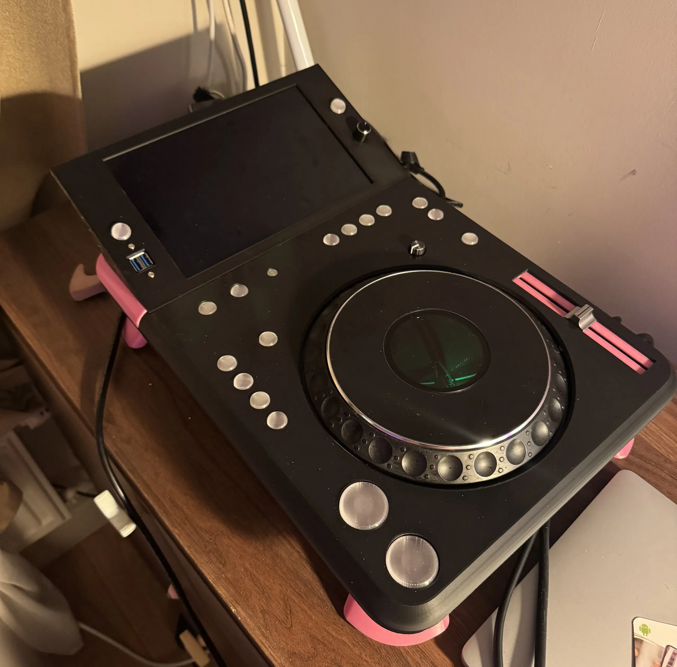 The deck seen from above at an angle on a wooden sideboard, screen off: a matte black printed top with rows of round silver buttons, a jog wheel glowing green at its centre, a tempo fader in a pink slot and a USB port beside the screen; pink feet lift it off the furniture.
