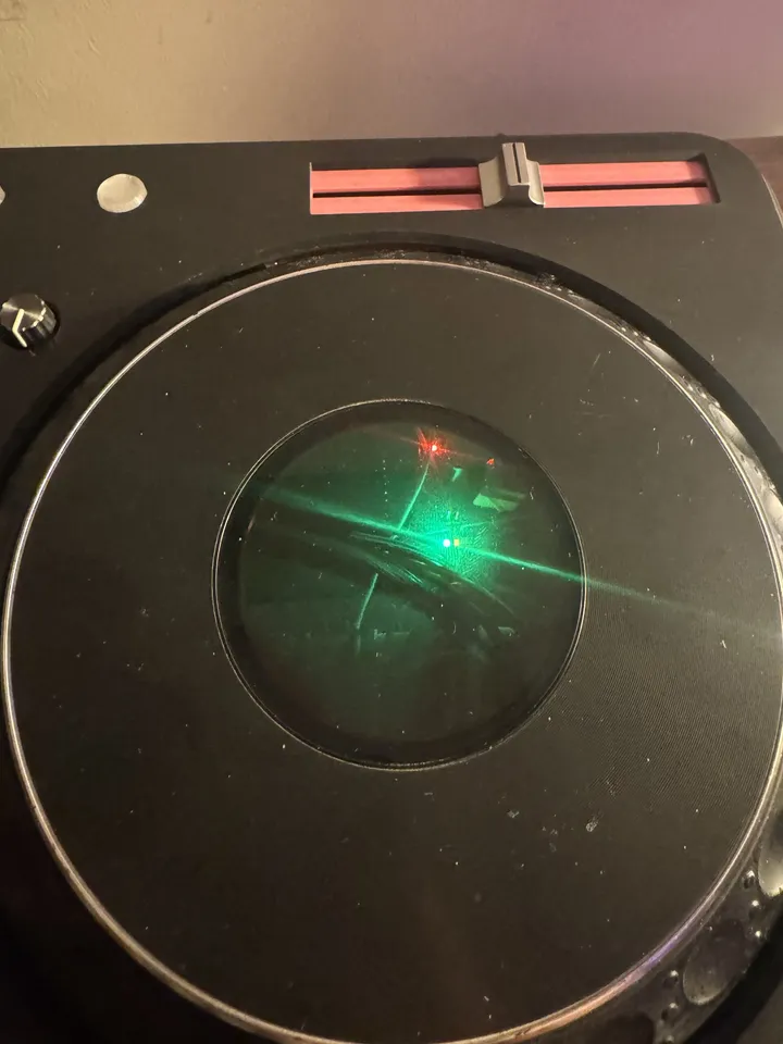 A top-down close-up of a black jog platter with a chrome rim; its translucent centre glows green with a red dot, and a pink tempo-fader slot sits above.