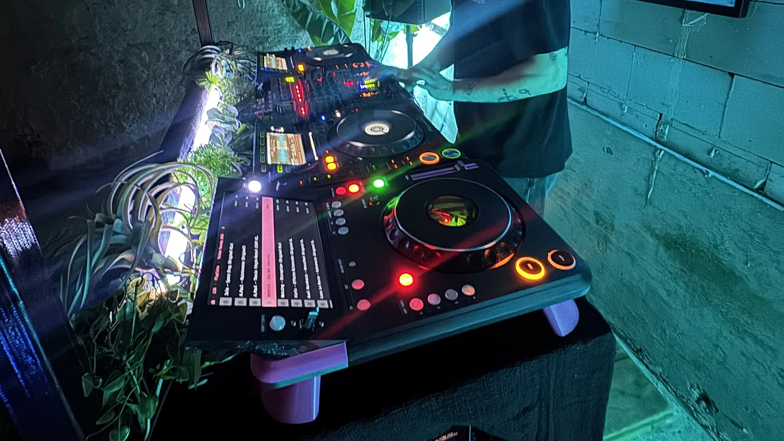 A teal-lit booth: the TriMixxx deck on pink legs in the foreground, its screen showing a pink-highlighted list, other equipment and a DJ's torso behind it, plants along the booth and a concrete-block wall.