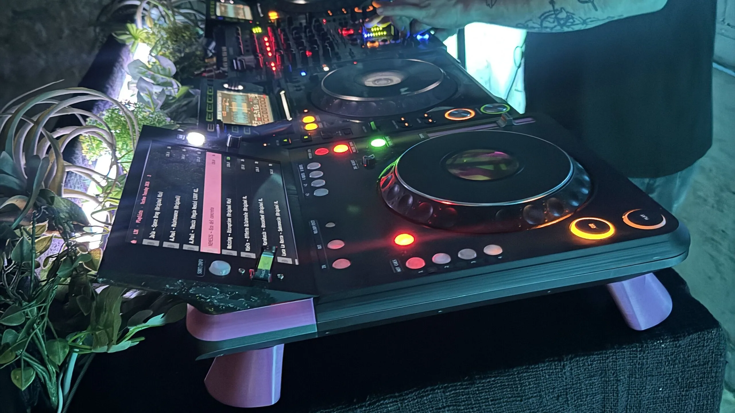A black DJ deck on pink legs, lit up in a dark booth: a tilted touchscreen showing a track list with one row highlighted in pink, a large jog wheel, and CUE and PLAY lit amber, with another player and a mixer behind it and a DJ's arm reaching past.