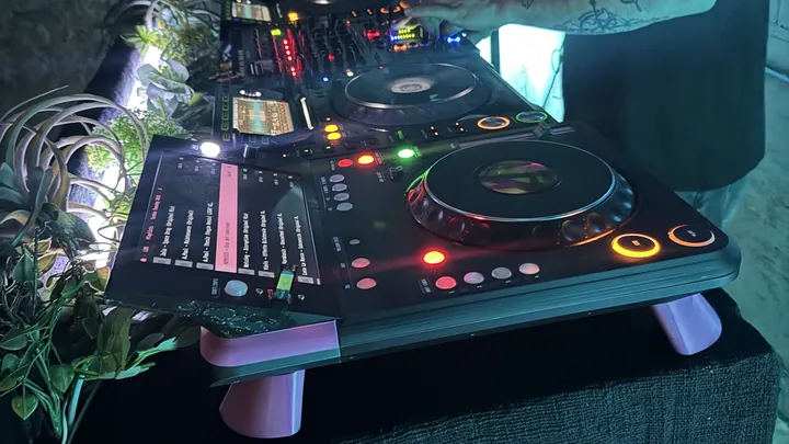 A black DJ deck on pink legs, lit up in a dark booth: a tilted touchscreen showing a track list with one row highlighted in pink, a large jog wheel, and CUE and PLAY lit amber, with another player and a mixer behind it and a DJ's arm reaching past.