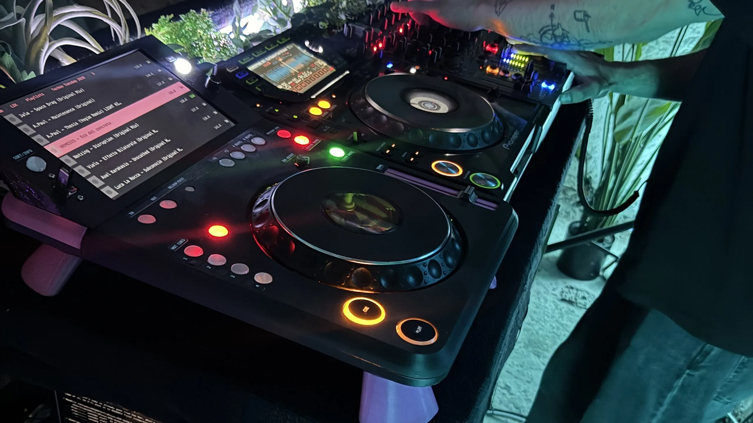 The deck in a dark booth from the front left: its touchscreen lists tracks with one highlighted in pink, the pads glow red and white and CUE and PLAY are lit amber; behind it another player and a mixer, worked by a DJ's tattooed arms.