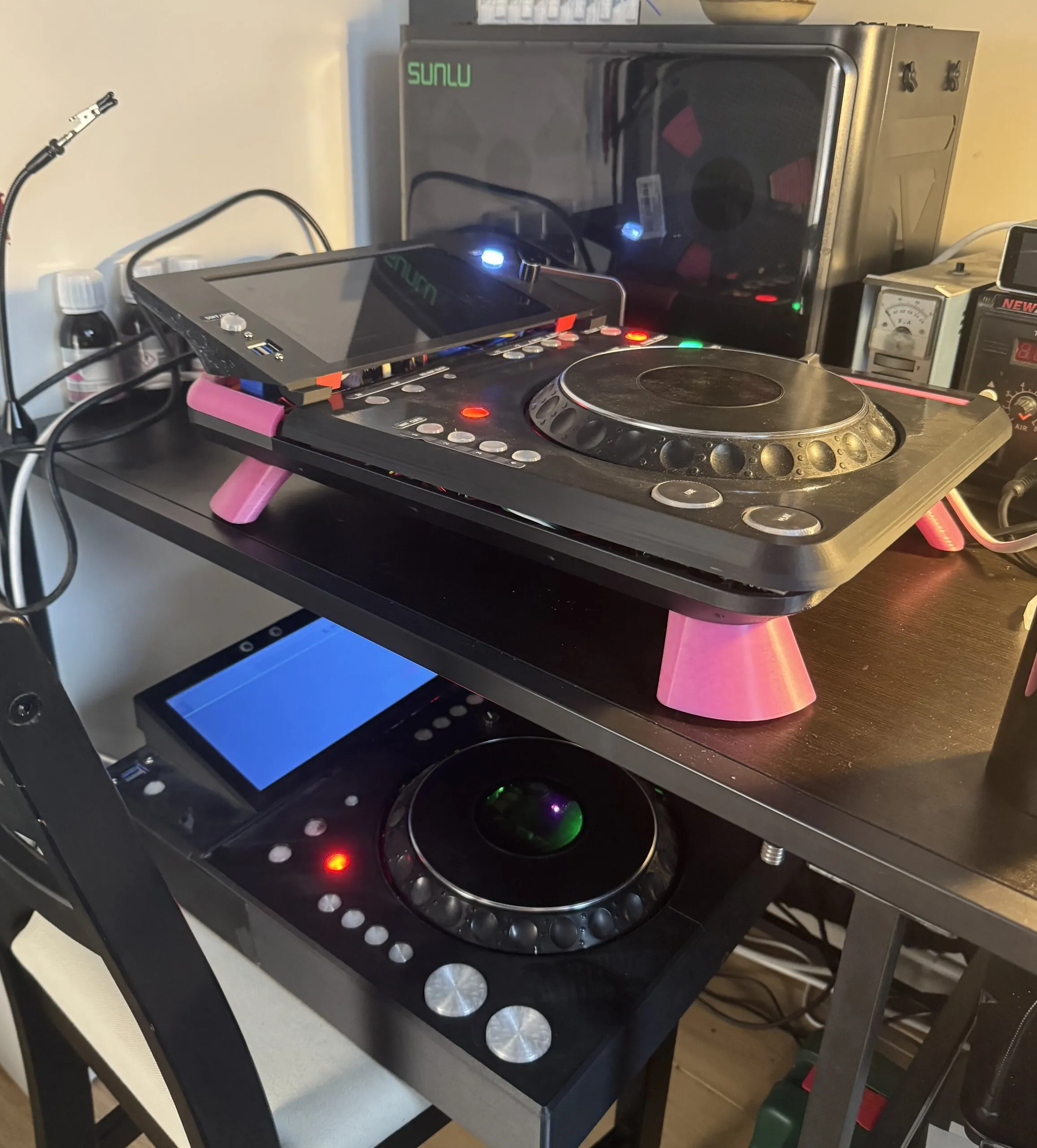 Two DJ decks at different heights: on a desk, the newer deck on pink cone-shaped legs with its tilted screen lit; below it, the older all-black deck with its screen glowing blue and its jog centre lit.