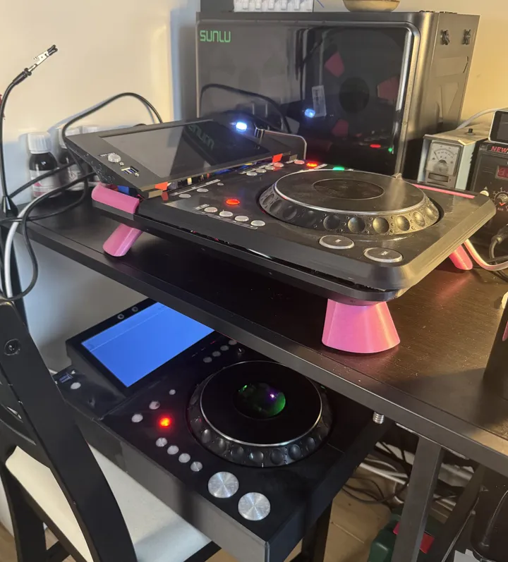 Two DJ decks at different heights: on a desk, the newer deck on pink cone-shaped legs with its tilted screen lit; below it, the older all-black deck with its screen glowing blue and its jog centre lit.